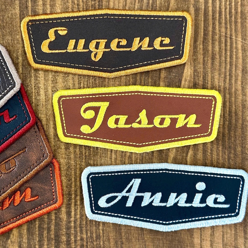 May include: A collection of personalized name patches. The patches are rectangular with a curved bottom edge. The names "Eugene", "Jason", and "Annie" are embroidered in a contrasting color on each patch. The patches have a stitched border.