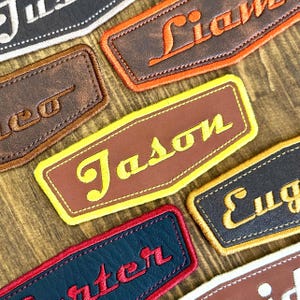 May include: Close-up of several personalized name patches. Each patch features a different name embroidered in a contrasting color on a leather-like material. The patches are in various colors, including brown, black, and blue, with names like "Jason" and "Liam".