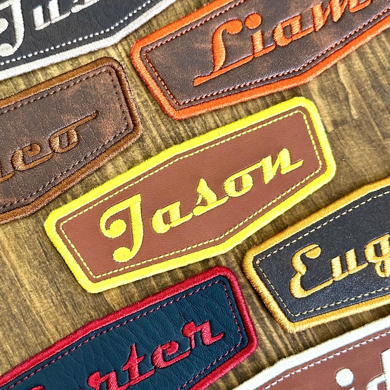 May include: Close-up of several personalized name patches. Each patch features a different name embroidered in a contrasting color on a leather-like material. The patches are in various colors, including brown, black, and blue, with names like "Jason" and "Liam".