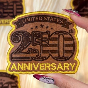 May include: A brown leather patch with gold stitching around the edges. The patch reads "UNITED STATES 250th ANNIVERSARY" in a vintage style. The number 250 has a stars and stripes design.