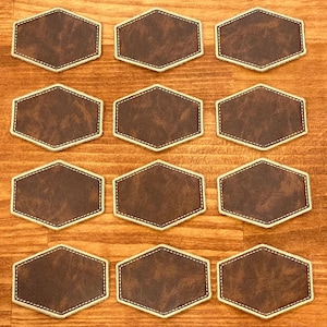 May include: Twelve brown leatherette coasters with a gold stitched border. The hexagonal coasters are arranged on a wooden surface. These coasters are a decorative item for the home.