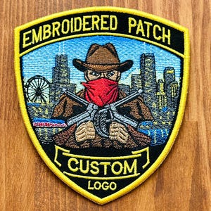 May include: Embroidered patch with a cowboy design. The patch features a cowboy with a red bandana, holding crossed weapons, and a cityscape background. The text "EMBROIDERED PATCH" and "CUSTOM LOGO" are displayed on the patch.