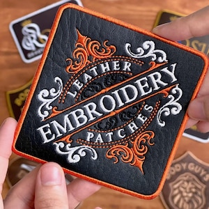 May include: Square black leather patch with orange trim and ornate white and orange embroidery. The patch reads "LEATHER EMBROIDERY PATCHES" in a decorative font. Other patches are visible in the background.