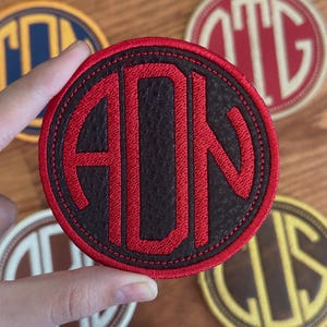 May include: A round, black and red monogram patch with the letters ADN in a bold, sans-serif font. The patch has a stitched border and is held by a hand. Other patches with different letter combinations are visible in the background.