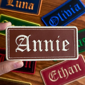 May include: A brown rectangular patch with the name "Annie" embroidered in white gothic lettering. The patch has a white stitched border. Other patches with names like "Luna", "Olivia", and "Ethan" are visible in the background.