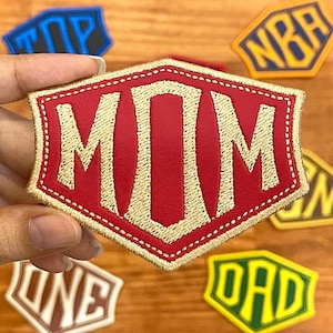 May include: A red patch with the word "MOM" embroidered in gold thread. The patch has a decorative border and is held by a hand. Other patches with the words "TOP", "NBA", "ONE", and "DAD" are also visible.