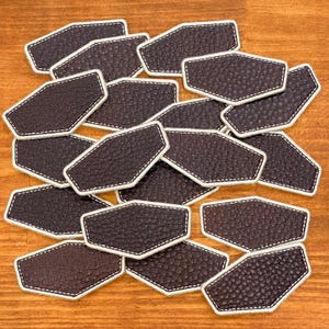 May include: A collection of brown leather patches with white stitching. Each patch has a unique shape with a textured surface. The patches are arranged on a wooden surface.