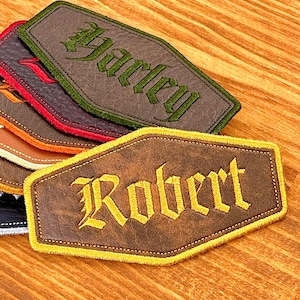 May include: A collection of personalized leather patches in various colors, each with a name embroidered in a gothic font. The patches are brown, green, red, orange, and black, with contrasting border stitching.