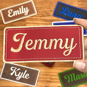 May include: A selection of name patches in different colours. The patches display names such as "Emily", "Luna", "Kyle", "Mason", and "Jemmy" in white or gold lettering. The rectangular patches have stitched borders.