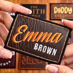 May include: A brown leather patch with the name "Emma" in orange script above the name "Brown" in white block letters. The patch has a black border. Other patches are visible in the background.