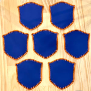 May include: Seven blue shield-shaped patches with orange trim and white stitching are arranged on a light wood surface. The patches have a smooth, solid blue center and a decorative border.