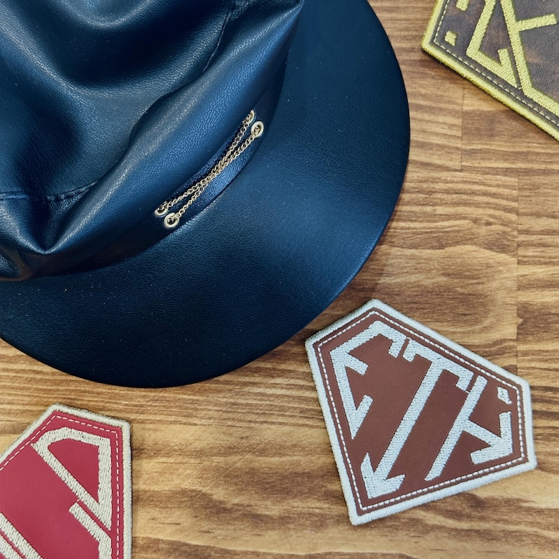 May include: A black leather hat with a gold chain detail and several embroidered patches. The patches are in the shape of a shield, with the letters "STA" and arrows. The patches are in red, brown, and yellow.