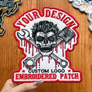 May include: Embroidered patch with a skull design, crossed wrenches, and a piston. The patch has a red border and features the text "YOUR DESIGN" and "CUSTOM LOGO EMBROIDERED PATCH". The skull has black hair and red blood dripping down.