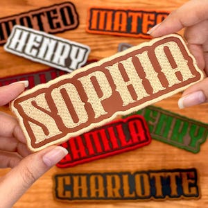 May include: A collection of embroidered name patches in various colors. The patches feature names like "Sophia," "Henry," and "Charlotte" in a Western-style font. The patches have a brown border and are displayed on a wooden surface.