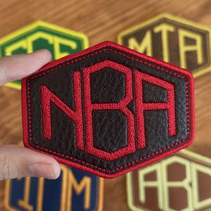 May include: A black and red embroidered patch with the letters "NBA" in red, stitched onto a black background. The patch is in a hexagonal shape. Other patches with different letter combinations are visible in the background.