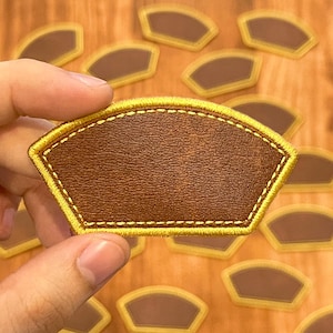 May include: A brown, fan-shaped patch with a textured surface and a yellow stitched border. The patch is held in a hand, with several similar patches visible in the background. The patches are likely for clothing or accessories.