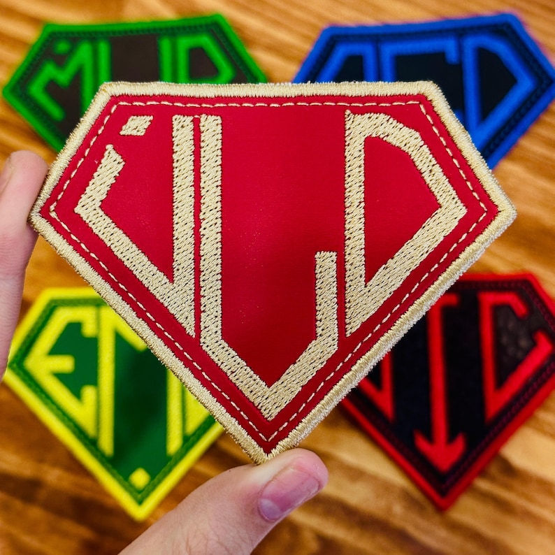 May include: A collection of embroidered patches in a diamond shape. The patches feature various color combinations, including red with gold stitching, green with white, and black with red. Each patch displays unique letter combinations.