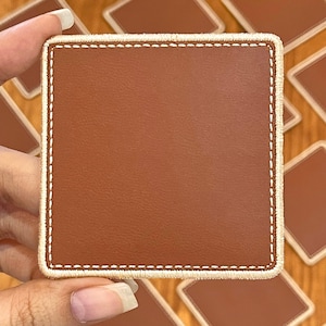 May include: A square brown coaster with a stitched white border. The coaster is held by a hand, with other coasters visible in the background. The coaster has a smooth, leather-like texture.