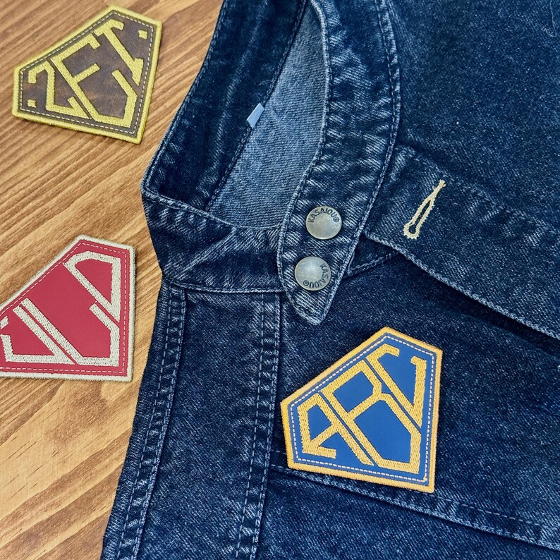 May include: Close-up of a dark blue denim jacket with three embroidered patches. The patches are diamond-shaped with the letters "ZET" and "RBV" in gold and blue. The jacket has silver buttons and a pocket.