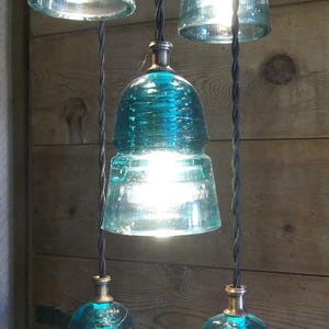May include: A cluster of pendant lights made from repurposed teal glass insulators. Each light fixture has a bulb inside, emitting a bright glow. The cords are black and twisted, and the background is a wooden wall.