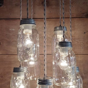 May include: A rustic chandelier made of six clear glass mason jars with metal lids, each with a light bulb inside. The jars are suspended from a metal ceiling fixture by gray braided cords.