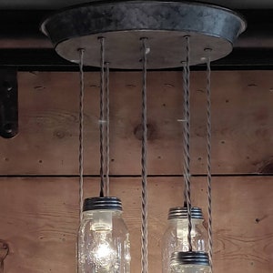May include: A rustic chandelier made from six clear glass mason jars with metal lids. The jars are suspended from a metal circular base by twisted cords.