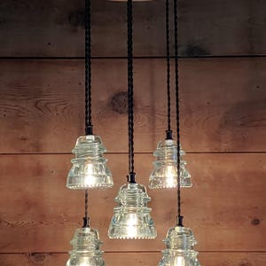 May include: A five-light pendant lamp with clear glass insulator shades. The shades hang from black cords attached to a circular fixture. The fixture is mounted on a wooden background. The light fixtures are illuminated.