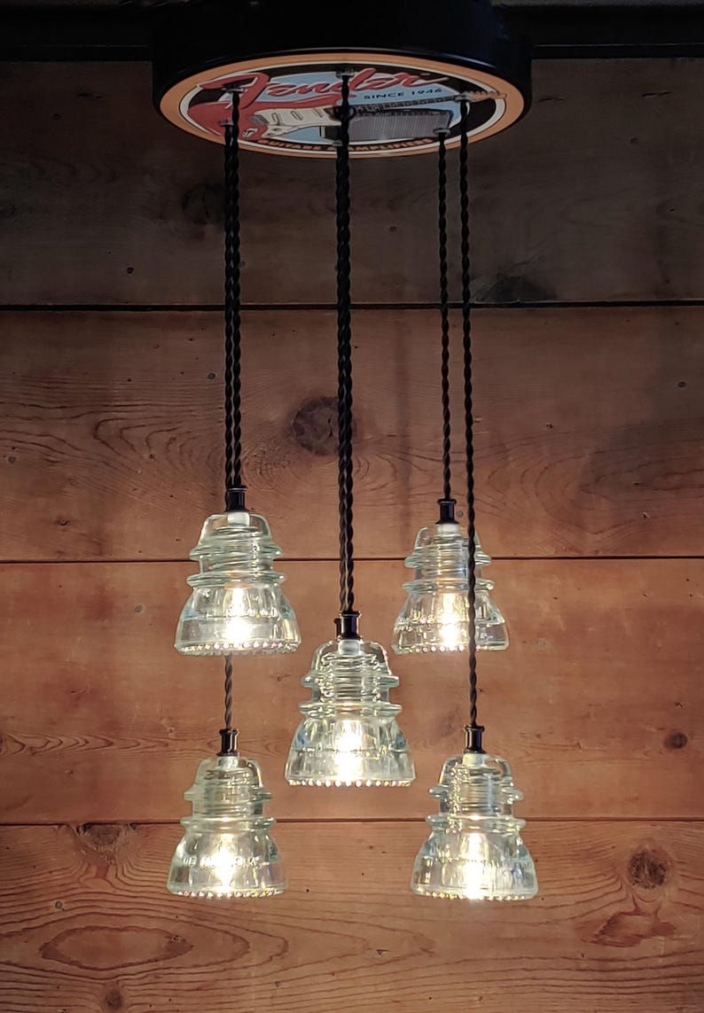 May include: A five-light pendant lamp with clear glass insulator shades. The shades hang from black cords attached to a circular fixture. The fixture is mounted on a wooden background. The light fixtures are illuminated.