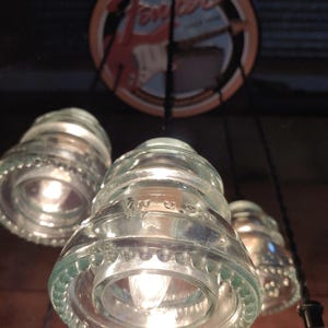 May include: A set of pendant lights made from recycled glass insulators, each with a warm light bulb. A circular sign with the Fender logo is visible above. The clear glass fixtures hang from black cords, creating a rustic, industrial style.