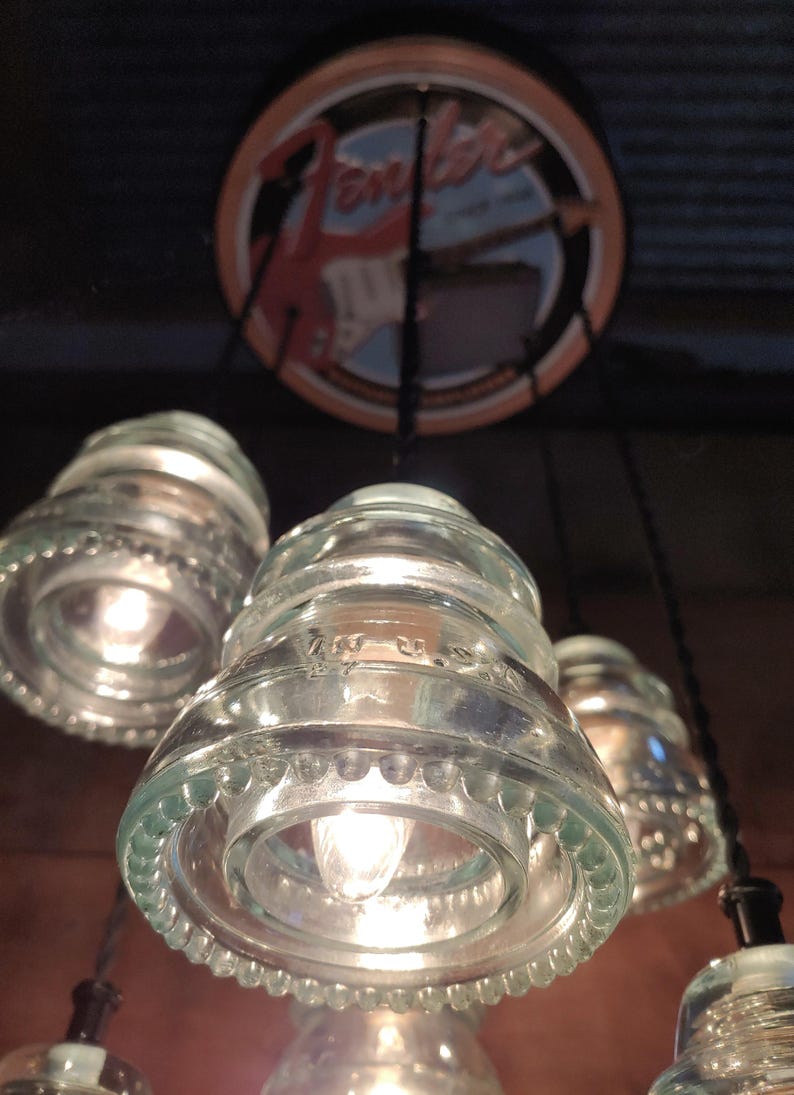 May include: A set of pendant lights made from recycled glass insulators, each with a warm light bulb. A circular sign with the Fender logo is visible above. The clear glass fixtures hang from black cords, creating a rustic, industrial style.