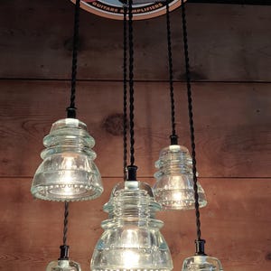 May include: A distinctive light fitting with five clear glass insulator pendant lights. The lights hang from black cords and are suspended from a Fender sign. The sign is round with a red and white guitar graphic and the words "Fender" and "Since 1946".