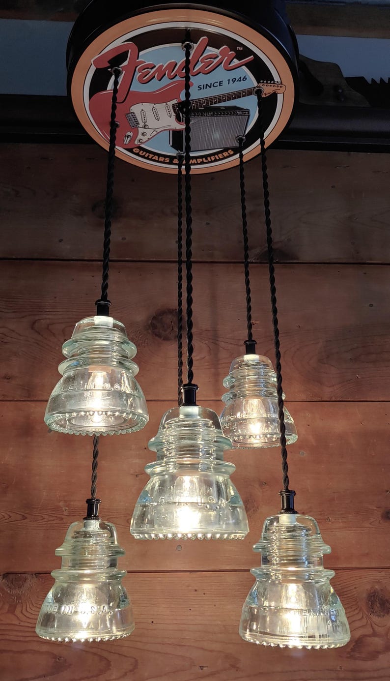 May include: A distinctive light fitting with five clear glass insulator pendant lights. The lights hang from black cords and are suspended from a Fender sign. The sign is round with a red and white guitar graphic and the words "Fender" and "Since 1946".