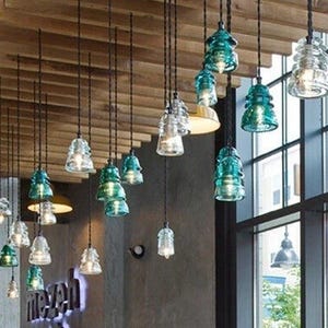 May include: A collection of hanging pendant lights with glass shades in clear and teal green. The lights are suspended from a wooden ceiling, creating a modern and stylish lighting design. The lights are in a restaurant or bar setting.