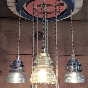 May include: A unique pendant light fixture featuring a wooden base with a blue and red Chicago Cubs logo. The fixture has multiple clear glass insulator lights hanging from twisted cords, creating a rustic and industrial aesthetic.