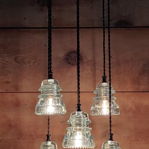 May include: A pendant light fitting featuring six clear glass insulator shades, each emitting a warm light. The shades are suspended from black cords attached to a circular fixture with the text "Fender" and "SINCE 1946". The backdrop is a wooden wall.