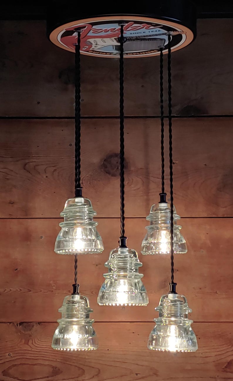 May include: A pendant light fitting featuring six clear glass insulator shades, each emitting a warm light. The shades are suspended from black cords attached to a circular fixture with the text "Fender" and "SINCE 1946". The backdrop is a wooden wall.