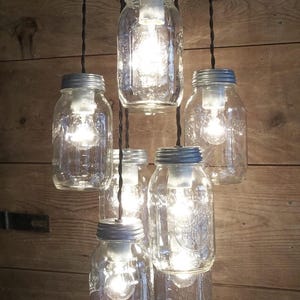 May include: A rustic chandelier made from clear glass mason jars with silver lids. Each jar houses a glowing light bulb, suspended from black cords. The fixture is set against a wooden plank background, creating a warm, inviting ambiance.