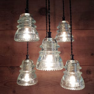 May include: Five clear glass pendant lights with Edison bulbs, each suspended by a black twisted cord. The lights have a vintage insulator design. The illuminated lights cast a warm glow against a wooden background, creating a rustic aesthetic.