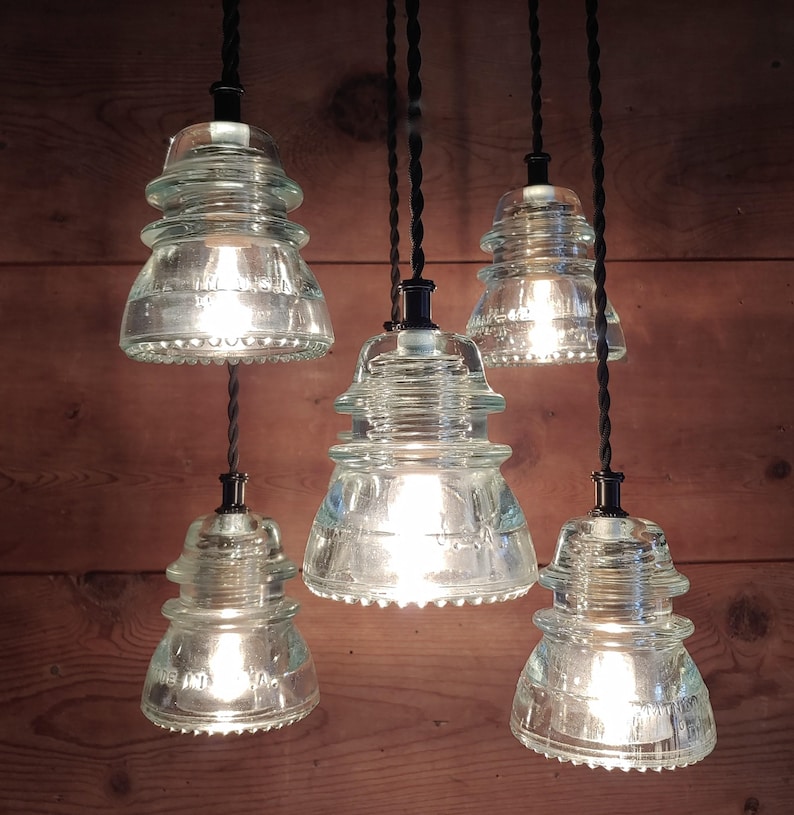 May include: Five clear glass pendant lights with Edison bulbs, each suspended by a black twisted cord. The lights have a vintage insulator design. The illuminated lights cast a warm glow against a wooden background, creating a rustic aesthetic.