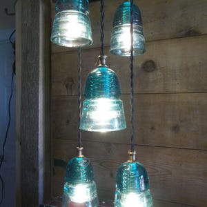 May include: A distinctive chandelier with five teal glass insulators. Each insulator holds a bright light bulb, suspended from black cords. The fixture hangs against a wooden wall, creating a rustic and industrial aesthetic.