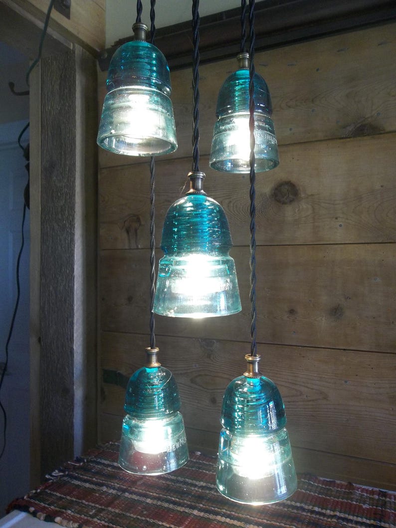 May include: A distinctive chandelier with five teal glass insulators. Each insulator holds a bright light bulb, suspended from black cords. The fixture hangs against a wooden wall, creating a rustic and industrial aesthetic.