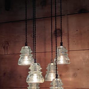 May include: A distinctive chandelier with six clear glass insulator light fittings hanging from a circular black base. The lights are suspended by black cords against a wooden backdrop. The fixture has a vintage, industrial style.