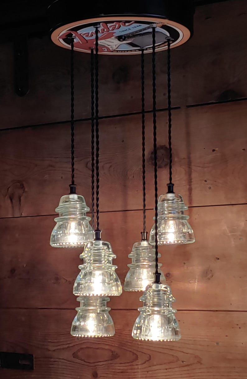 May include: A distinctive chandelier with six clear glass insulator light fittings hanging from a circular black base. The lights are suspended by black cords against a wooden backdrop. The fixture has a vintage, industrial style.