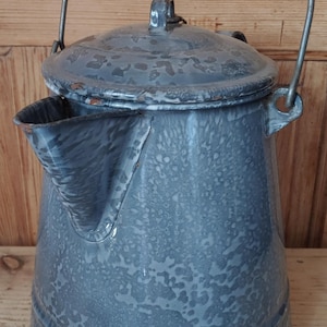 May include: A vintage gray enamel coffee pot with a speckled finish, featuring a wide spout, a matching lid with a small handle, and a metal swing handle with a black wooden grip. The surface shows signs of wear and rust along the edges.