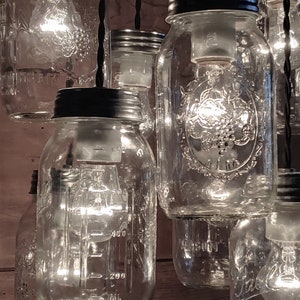 May include: A group of clear glass mason jars with black lids are hanging from black cords. Each jar has a light bulb inside, and the jars are illuminated. The jars are arranged in a cluster, with some overlapping. The jars are all the same size and shape, and they have a simple, rustic design.