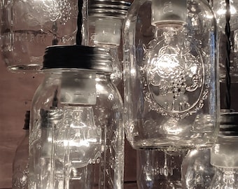 May include: A group of clear glass mason jars with black lids are hanging from black cords. Each jar has a light bulb inside, and the jars are illuminated. The jars are arranged in a cluster, with some overlapping. The jars are all the same size and shape, and they have a simple, rustic design.