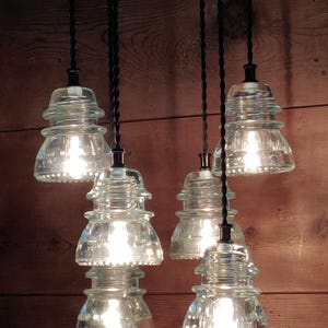 May include: Six clear glass pendant lights, styled like old insulators, suspended from black cords. The lights are illuminated, creating a warm glow against a wooden backdrop. The lights are a unique home decor item.