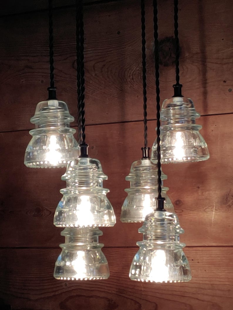 May include: Six clear glass pendant lights, styled like old insulators, suspended from black cords. The lights are illuminated, creating a warm glow against a wooden backdrop. The lights are a unique home decor item.