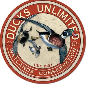May include: Circular Ducks Unlimited Wetlands Conservation emblem. The design features three ducks in flight against a backdrop of a wetland scene. The emblem is red with white text and a cream-colored inner circle. The text reads "DUCKS UNLIMITED" and "WETLANDS CONSERVATION EST. 1937".