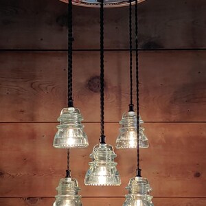 May include: A distinctive light fitting with six clear glass insulator pendant lights hanging from a circular fixture. The fixture has a vintage look, with black cords and a wooden background. The top fixture has the text "Fender Since 1946".